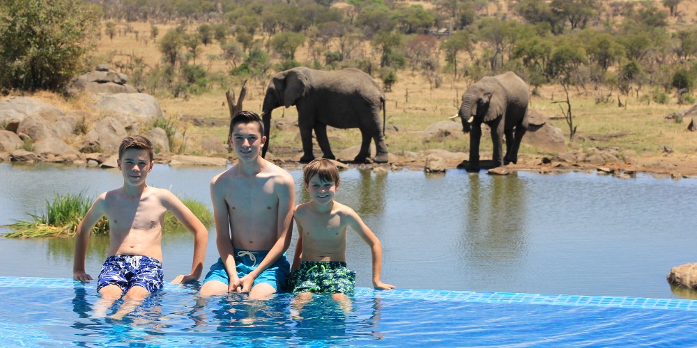 three-boys-sitting-on-edge-of-infinity-pool-with-elephants-in-background-tanzania-safari