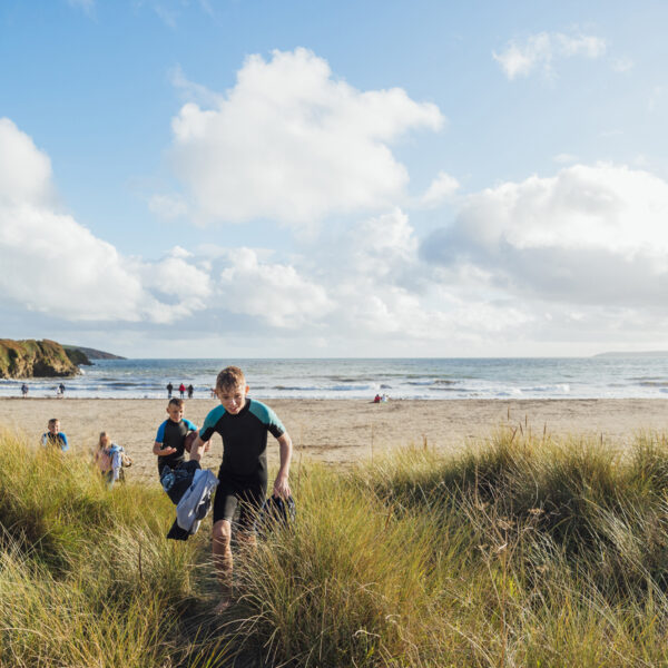 Boys wearing wetsuits walking from the beach on a sunny day in autumn. They are on a staycation with their family to lower their carbon footprint and support the local economy.