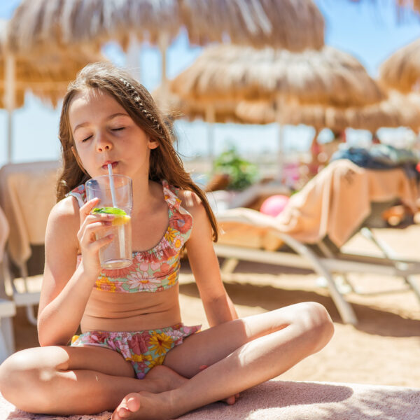 Young girl in floral bikini drinking refreshing non-alcoholic mojito through straw while sitting cross-legged on beach towel under sun umbrellas